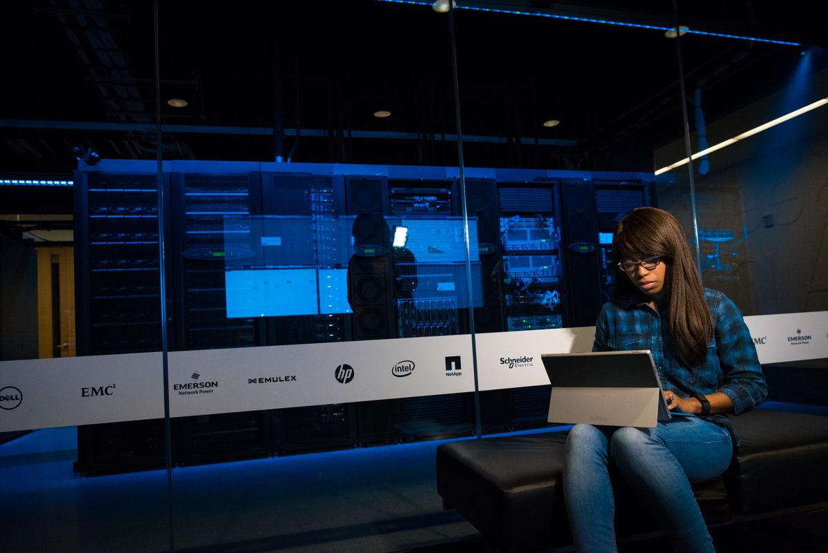 Woman with a tablet sitting in front of a glass wall and blue-lit server racks with vendor logos